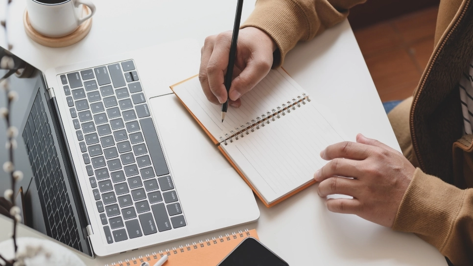 Egy asztalon laptop és vonalas jegyzetfüzet látható. Egy férfi két keze az asztalon van, az egyikben ceruzát fog és a füzetbe készül írni. A széleken részben egy bögre kávé és egy mobiltelefon is látszódik.