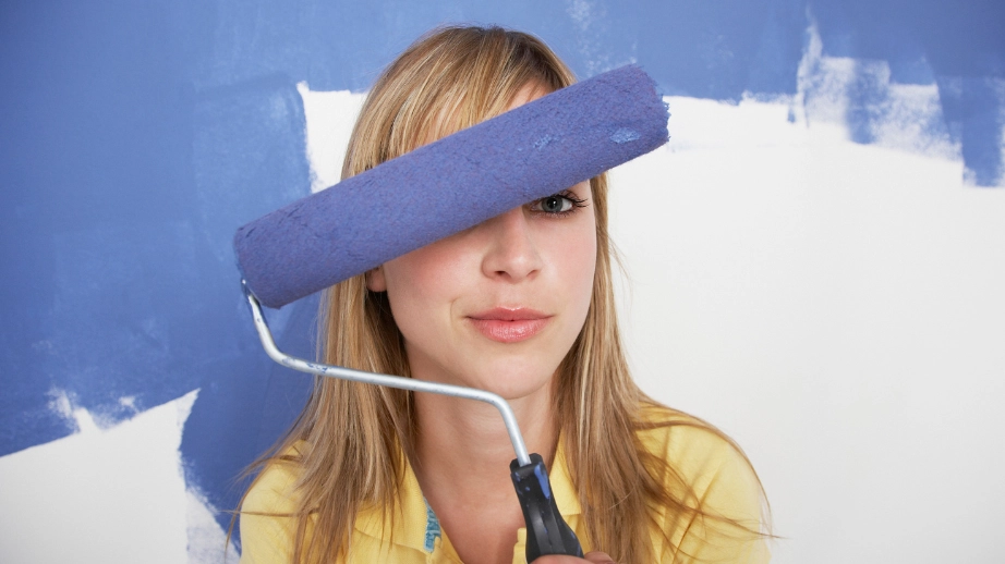 Nő házilagos festésmunkát végez, kék színű festékhengerrel félkész fehér falat fest.