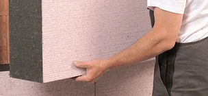 A képen egy férfi vastag hőszigetelő anyaggal von be egy falat. A szakembernek csak a keze látszik, fehér pólót és szürke nadrágot visel.