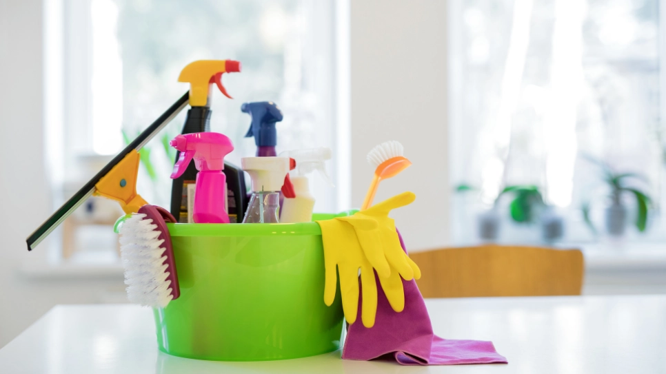 Különböző tisztítószerek egy zöld vödörben. A tisztítószereken kívül más eszközök, ablakpucoló, kefe, gumikesztyű, és mikroszálas kendő látszódnak.