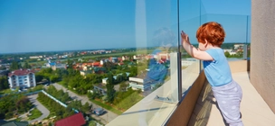 Csecsemő támaszkodik üvegkorlátnak egy magas épület erkélyén.